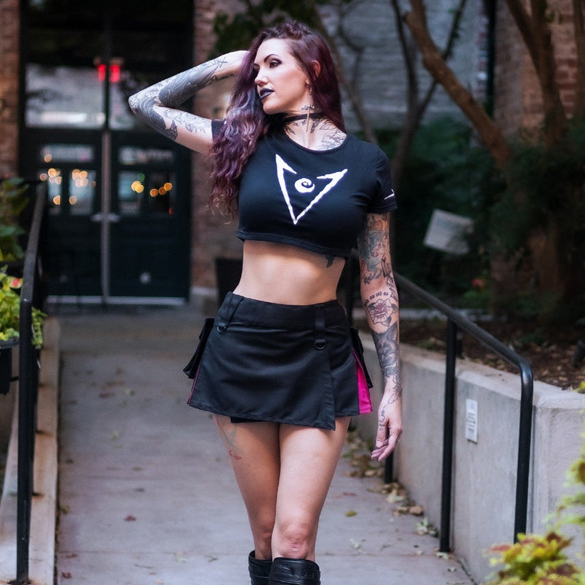 A woman with tattoos models a black crop top and kilt, paired with black lace-up boots, walking on an outdoor concrete pathway.