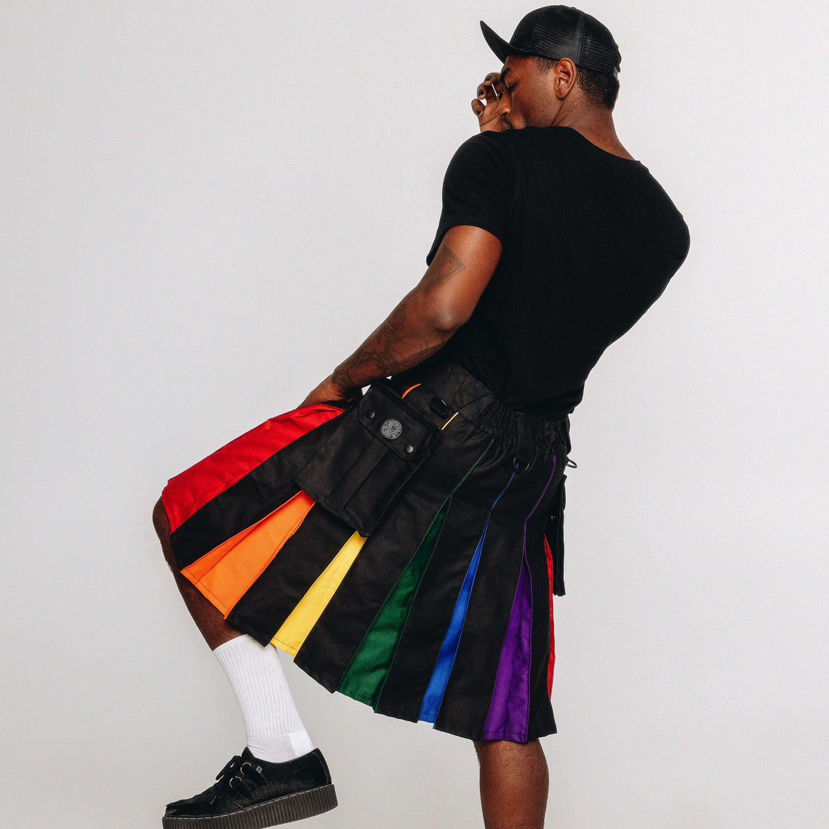 A man wearing a black cap, black t-shirt, white socks, and black platform shoes models a black pleated kilt with vibrant rainbow-colored fabric panels. He is photographed from behind against a solid light gray studio background.