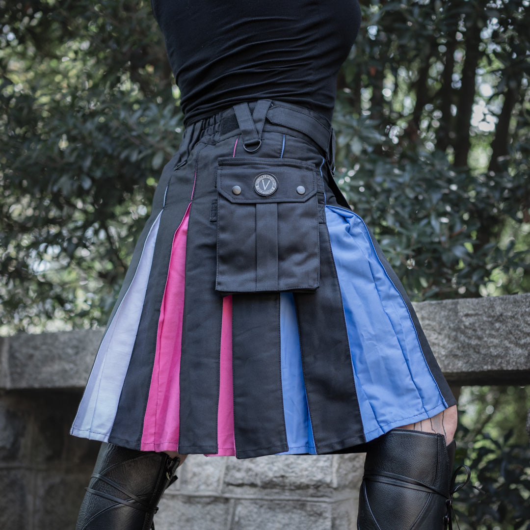 A person outdoors wearing a black pleated kilt-style skirt with vertical color-block panels in white, pink, black, blue, and light blue, plus a front flap pocket. Trees and a stone wall are visible in the background.