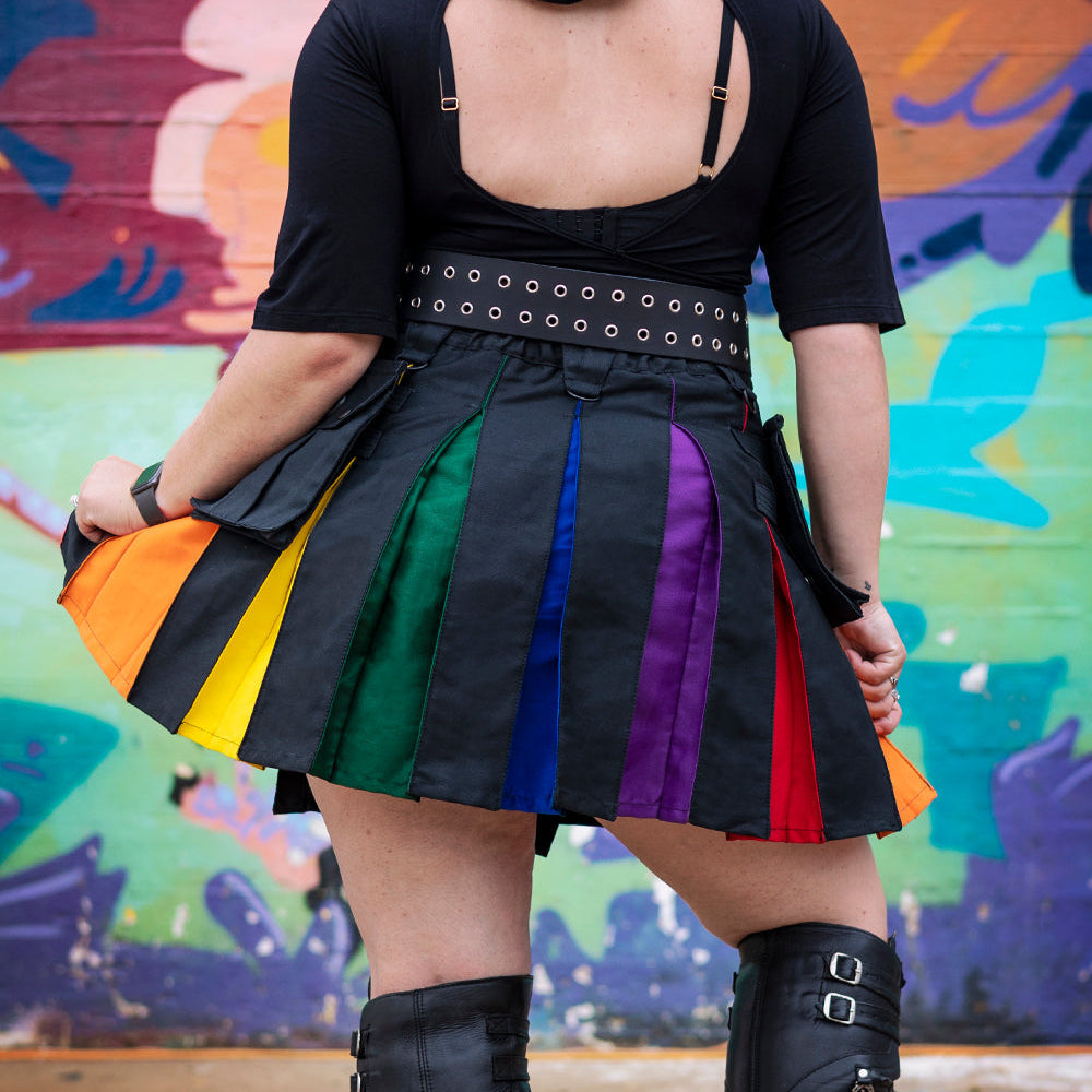 A person shown from the back wearing a black short-sleeve top with an open back, a black pleated kilt with vertical rainbow-colored panels, and black knee-high lace-up boots, standing in front of a colorful graffiti wall.