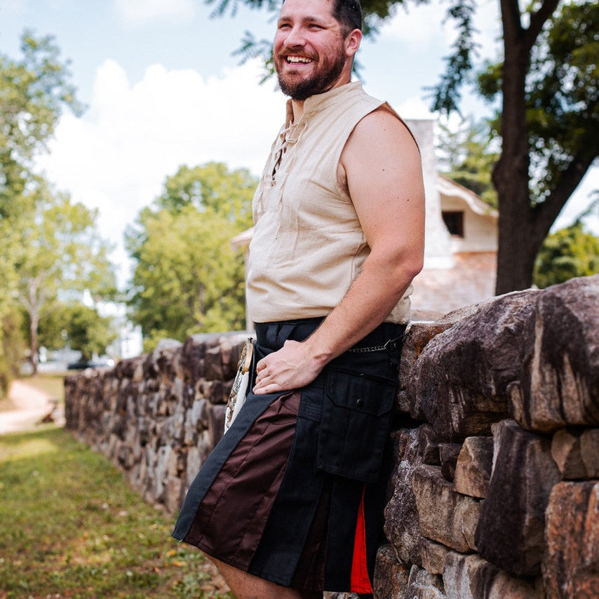 A man wearing a gay bear pride inspired kilt with black, brown, and orange toned panels, leaning against a rustic stone wall outdoors.