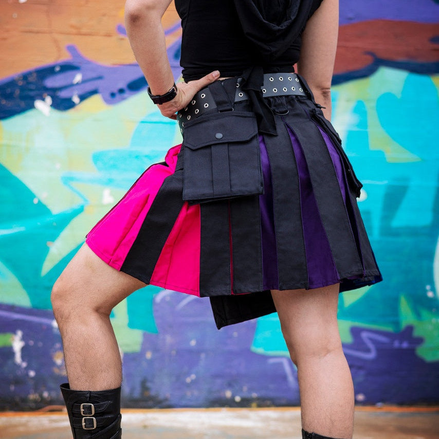 A person wearing a black goth-style pleated mini kilt with bright pink and purple panels, black knee-high boots, and a black top, standing in front of a colorful graffiti wall.