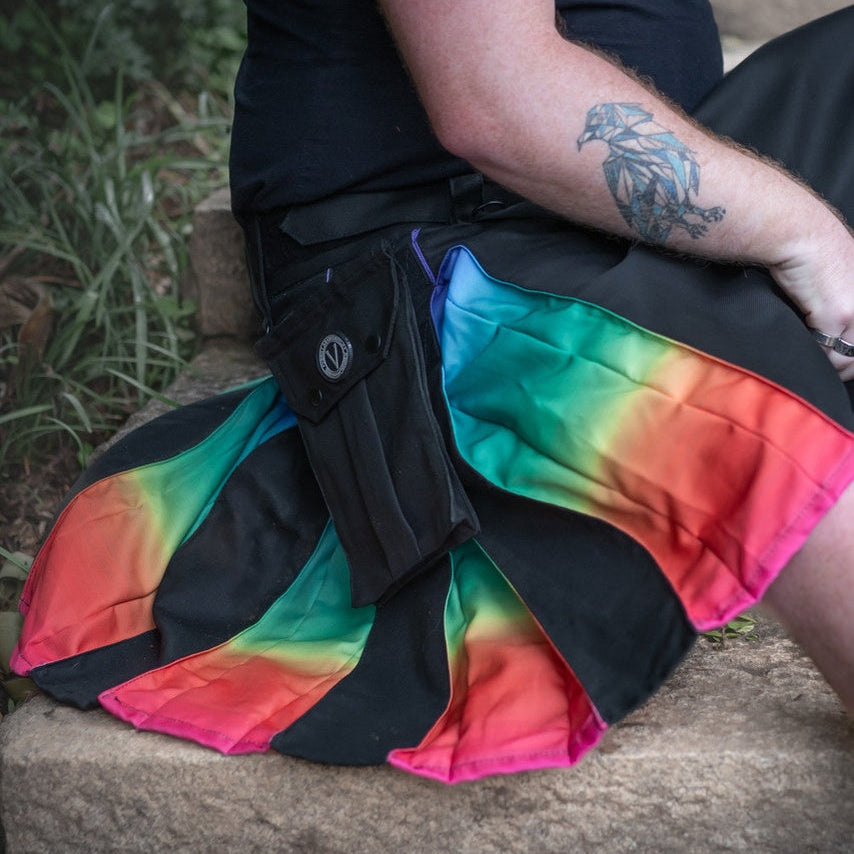 A person sitting outdoors on stone steps wearing a black kilt with a vibrant rainbow gradient pattern and a black utility pouch at the hip.