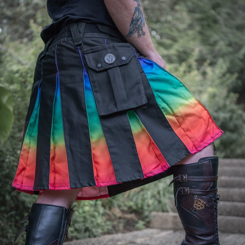 A person outdoors wearing a black pleated kilt with vertical rainbow gradient panels in blue, green, yellow, orange, and pink, paired with black knee-high boots.